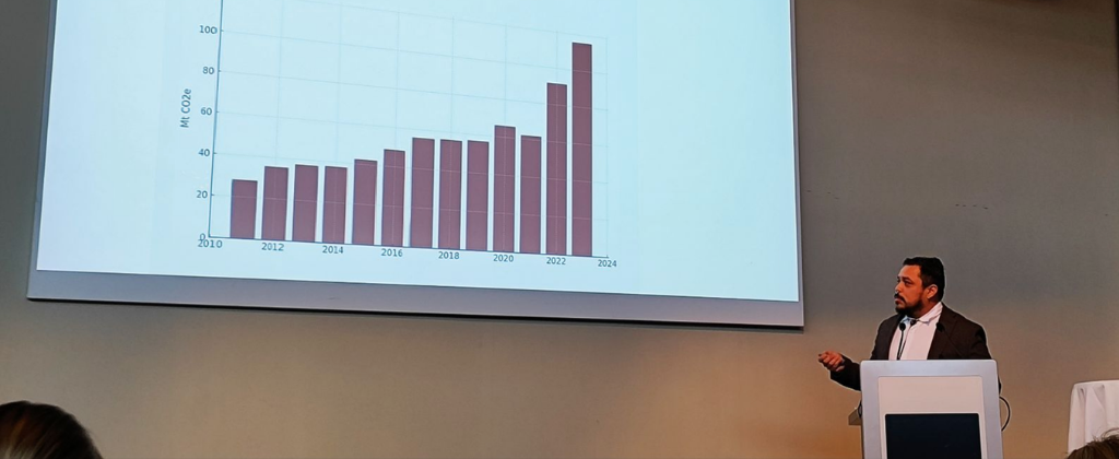 Guido Núñez-Mujica at a lectern at European Nuclear Energy 2026, gesturing toward a slide charting rising Mt CO2e from 2010 to 2024