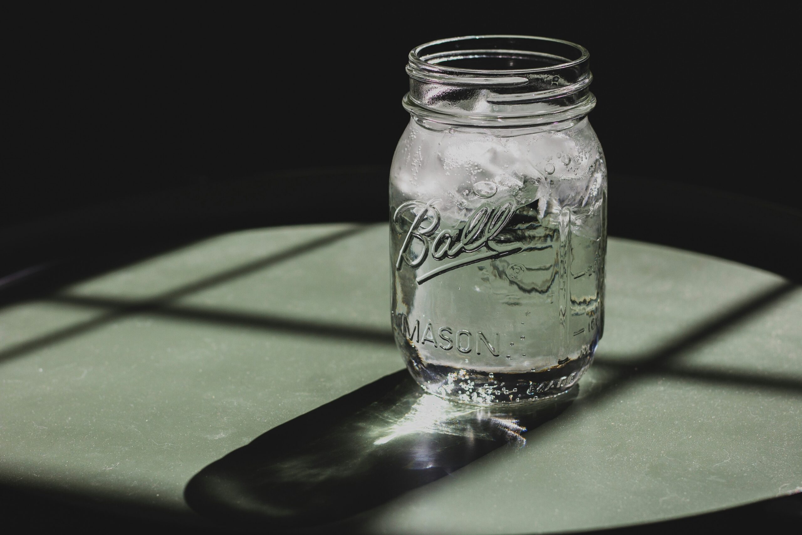 A Ball mason jar of clear water with ice, lit by hard sunlight on a green table