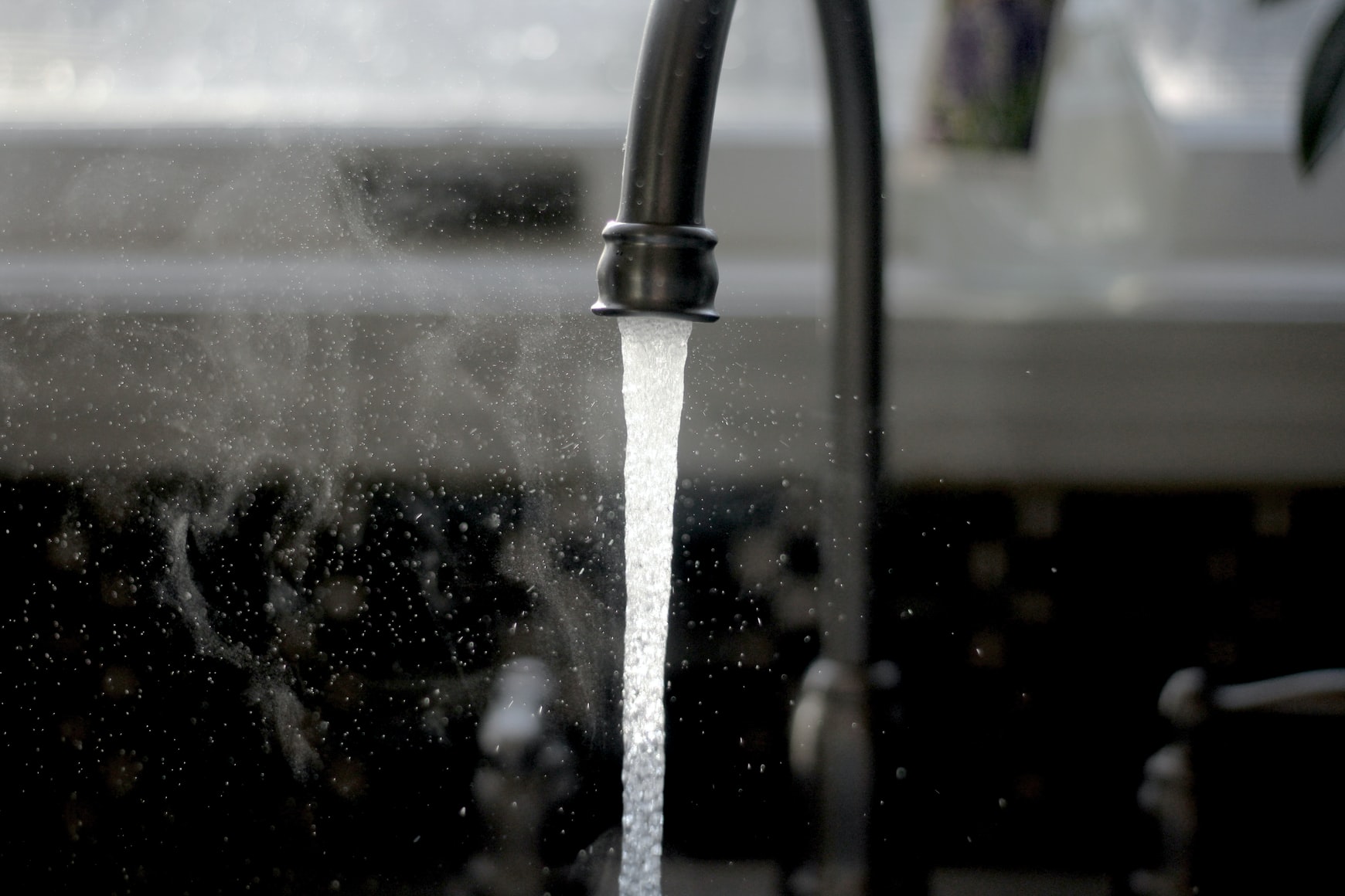 Water running from a dark kitchen faucet into a sink, droplets suspended in the light