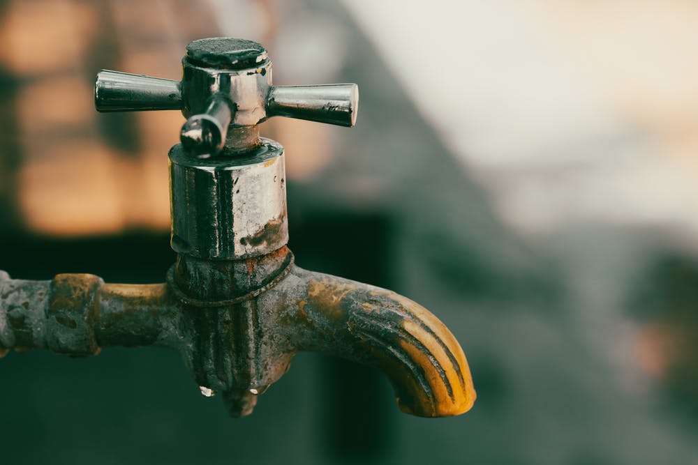 Close-up of a weathered outdoor tap, corroded and stained, with a drop of water forming at the spout