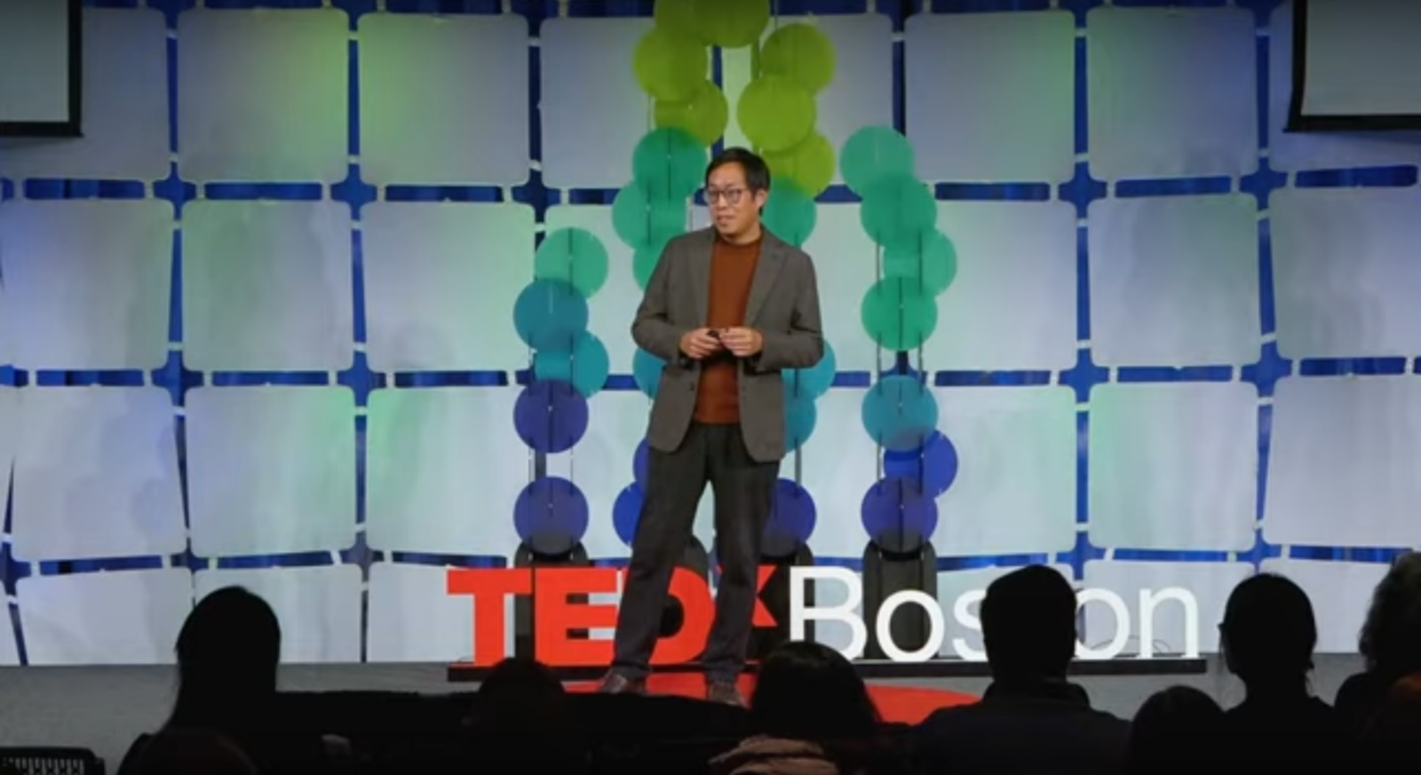 Dr. Frank Ling standing on the TEDxBoston stage in front of a tiled backdrop and hanging green and blue discs